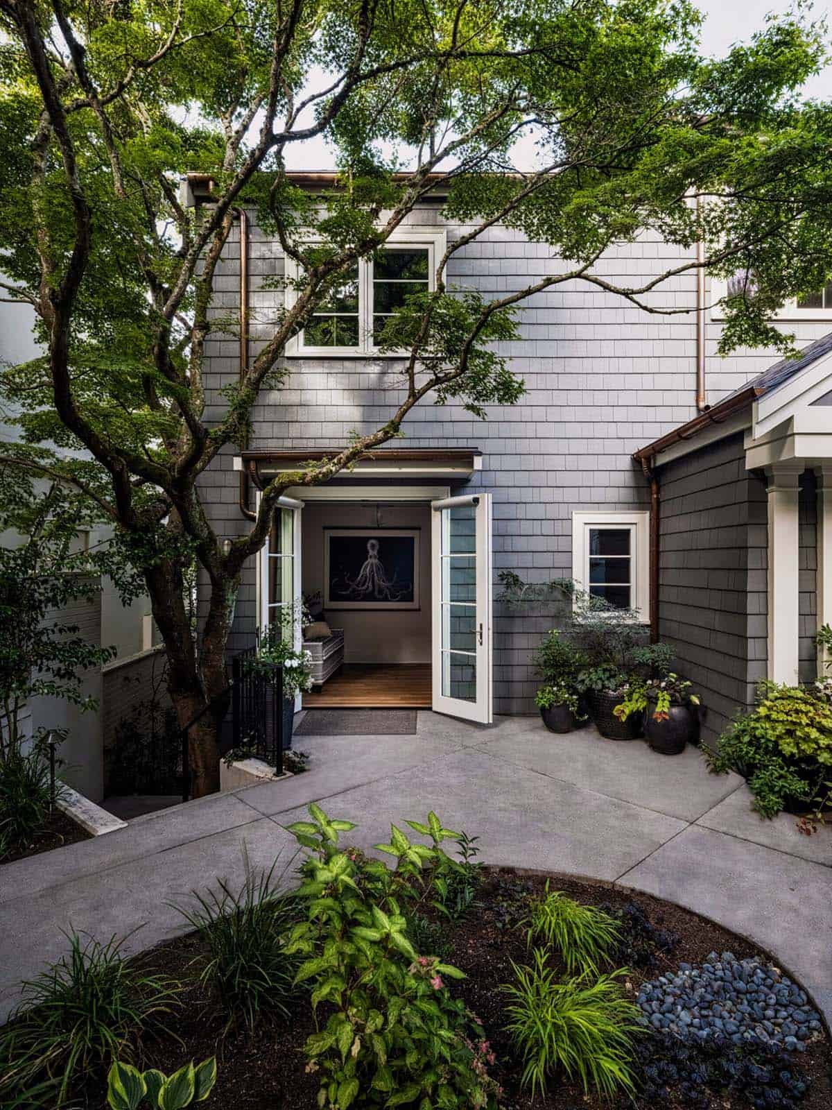 Side entrance of gray shingle home with open French door revealing octopus artwork and mature Japanese maple tree