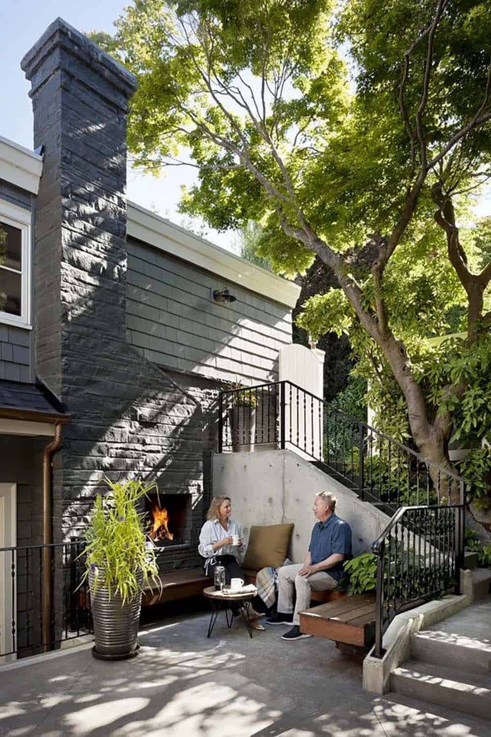 Couple relaxing beside outdoor stone fireplace on wood bench patio with tree canopy overhead