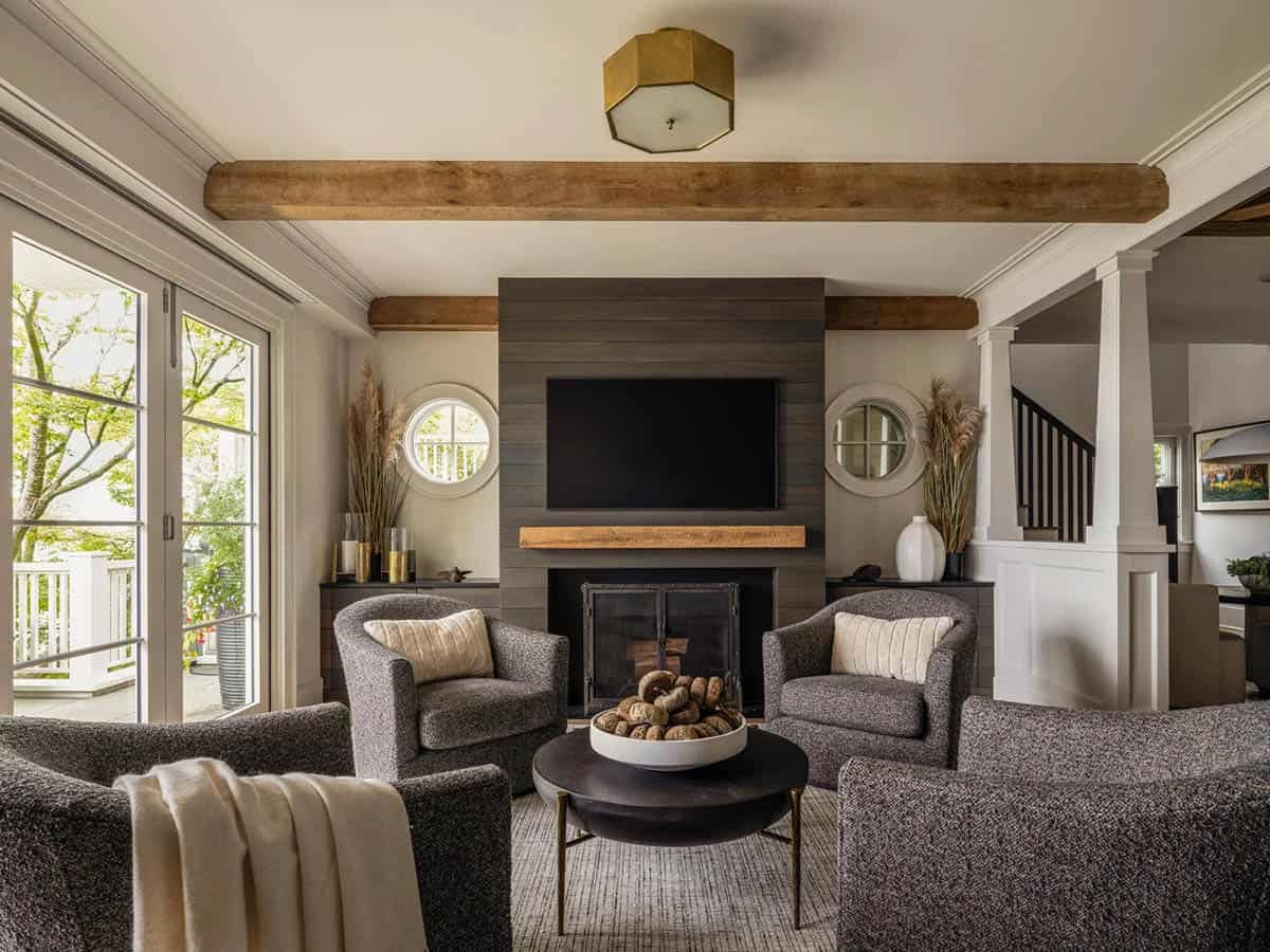 Living room with shiplap fireplace wall, flanking porthole windows, gray armchairs, and exposed wood beam