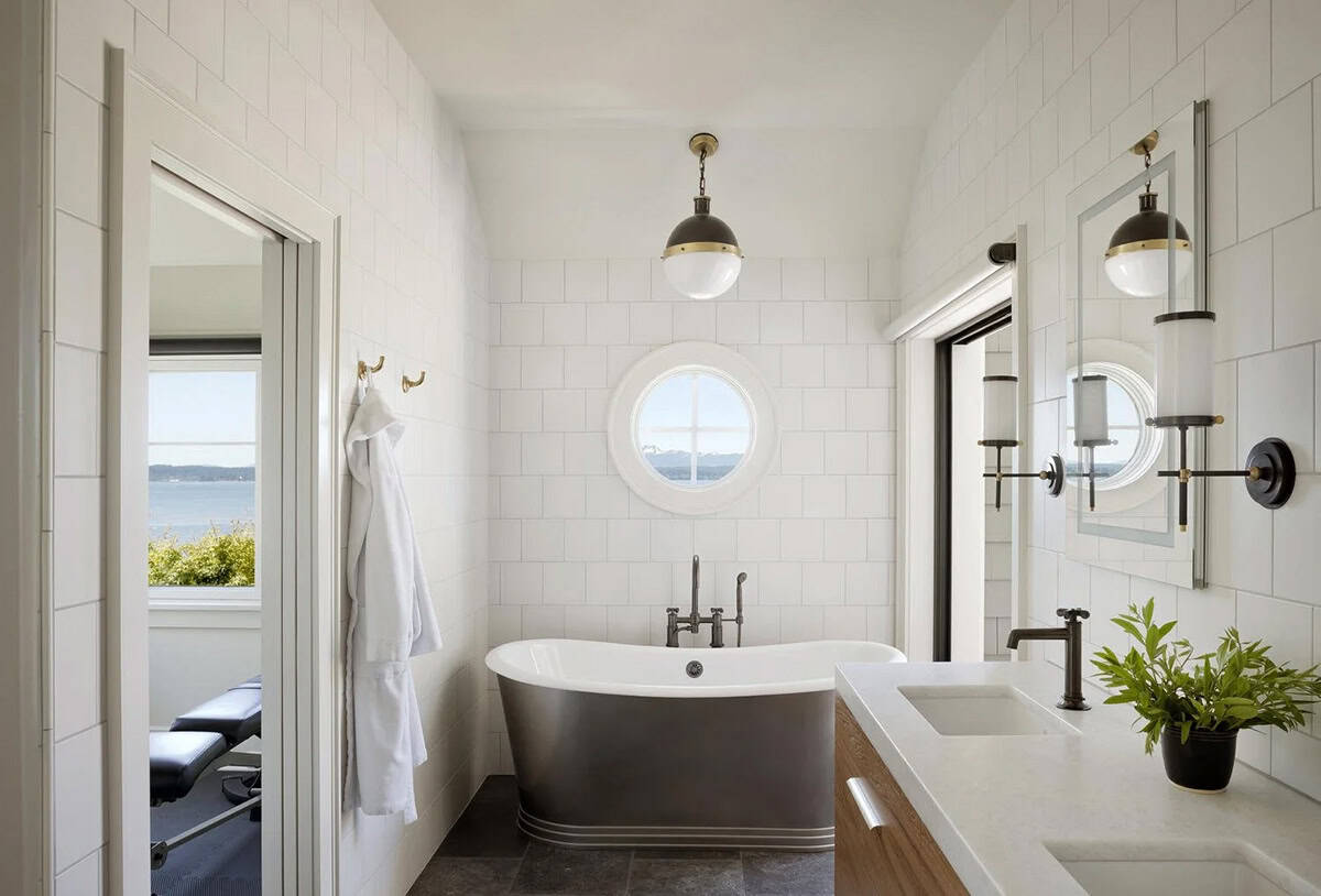 Primary bathroom with freestanding dark soaking tub, porthole window, white tile walls, and brass pendant