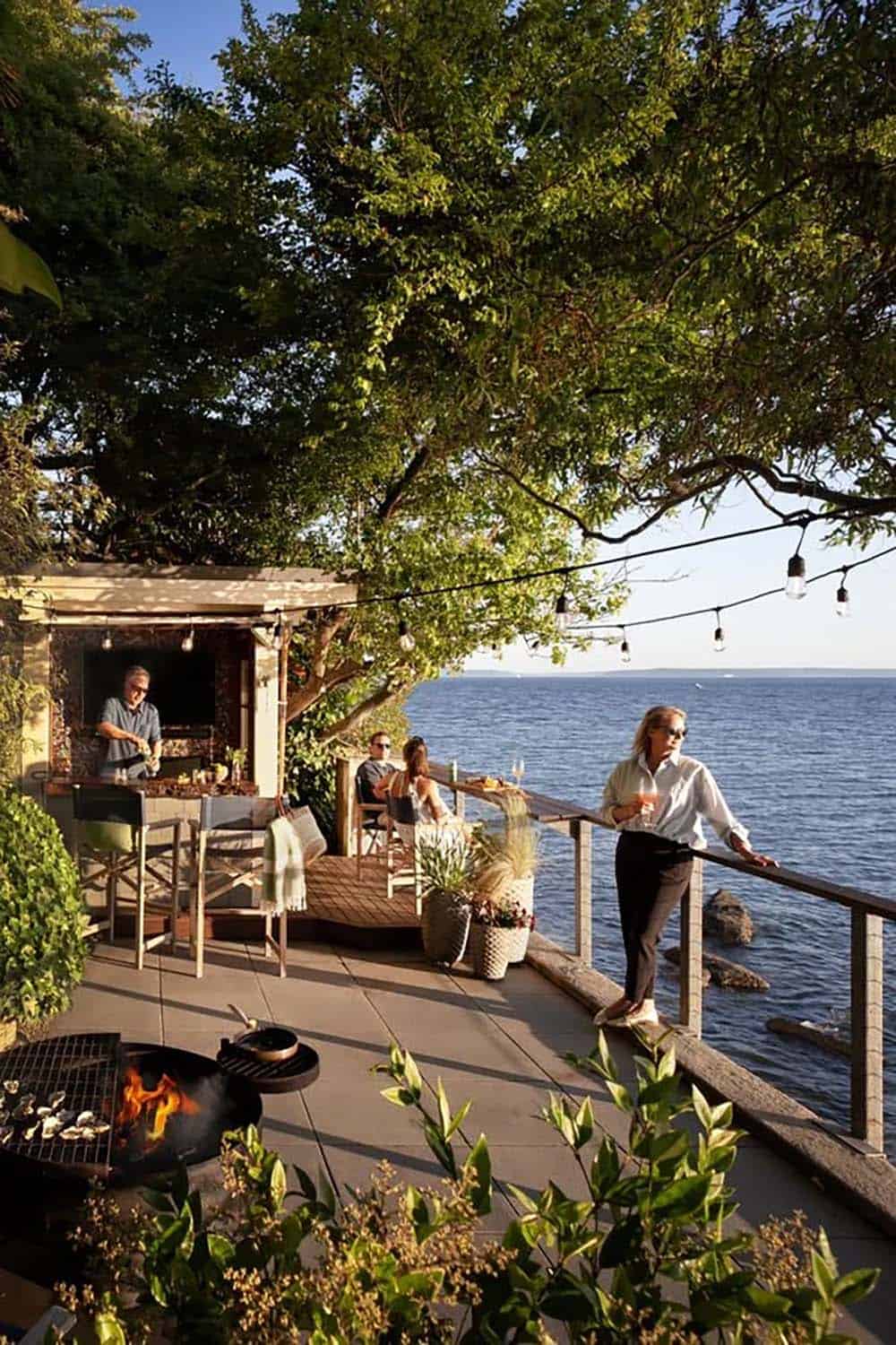 Group gathering on waterfront terrace at sunset with string lights, fire pit, and original boathouse bar structure