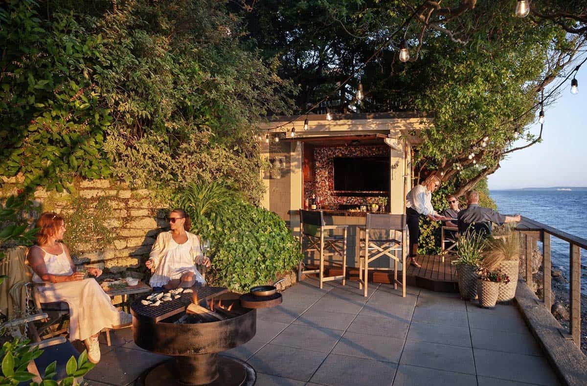 Evening entertaining on waterfront stone terrace with fire pit, string lights, lush greenery, and original boathouse bar