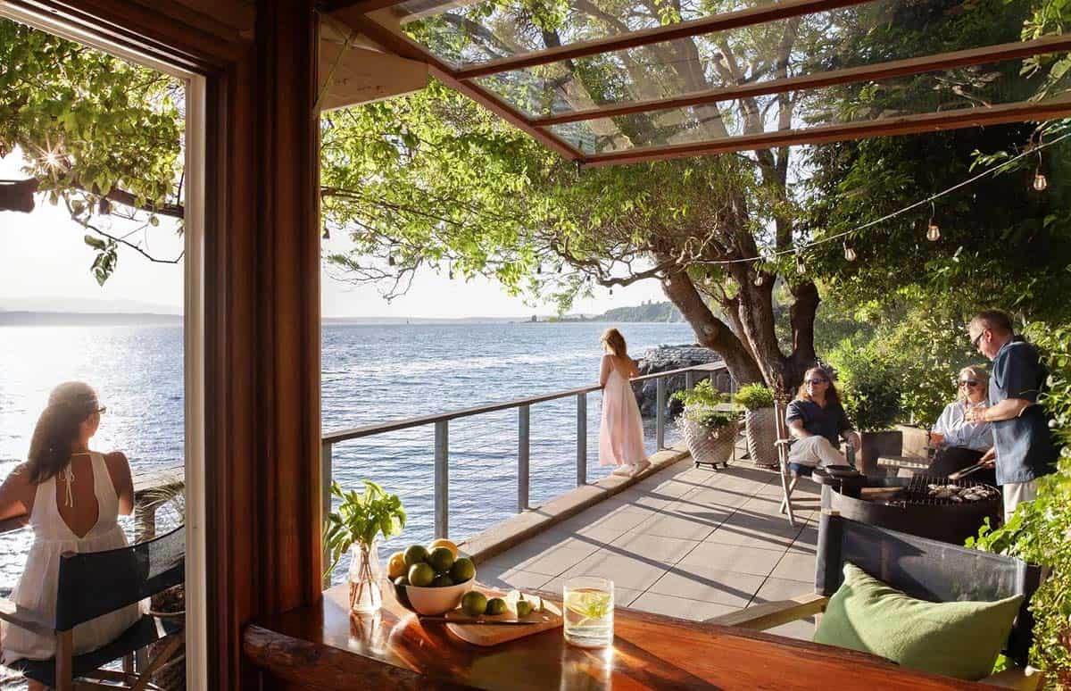 View from boathouse interior through glass roof pergola to waterfront terrace with group socializing at sunset