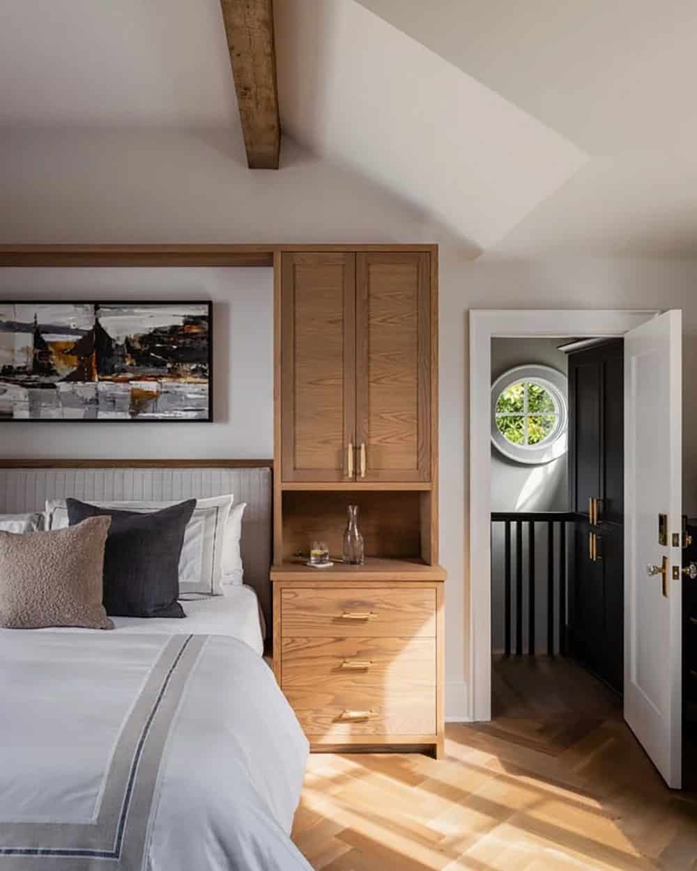 Primary bedroom detail showing oak built-in nightstand and wardrobe with brass hardware, herringbone floor, and porthole window beyond