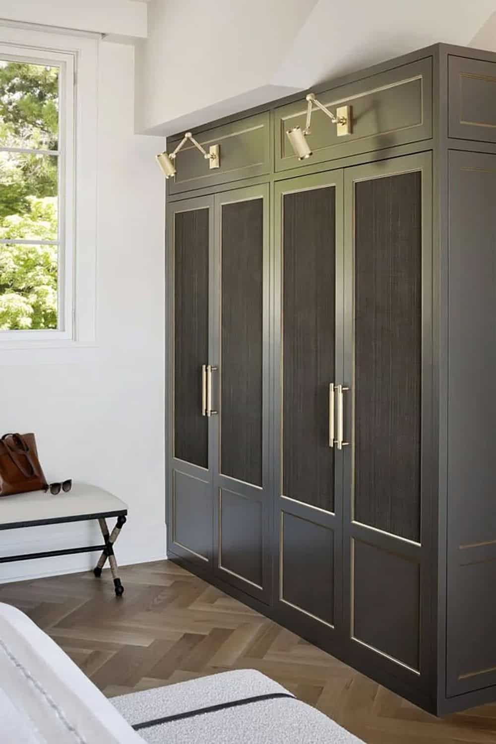 Dark charcoal built-in wardrobe with grasscloth panel doors, brass hardware, and brass picture lights on herringbone floor