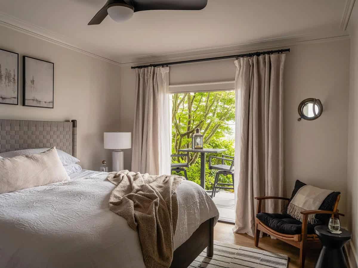 Guest bedroom with woven headboard, linen drapes, wood chair, and garden balcony view through French door