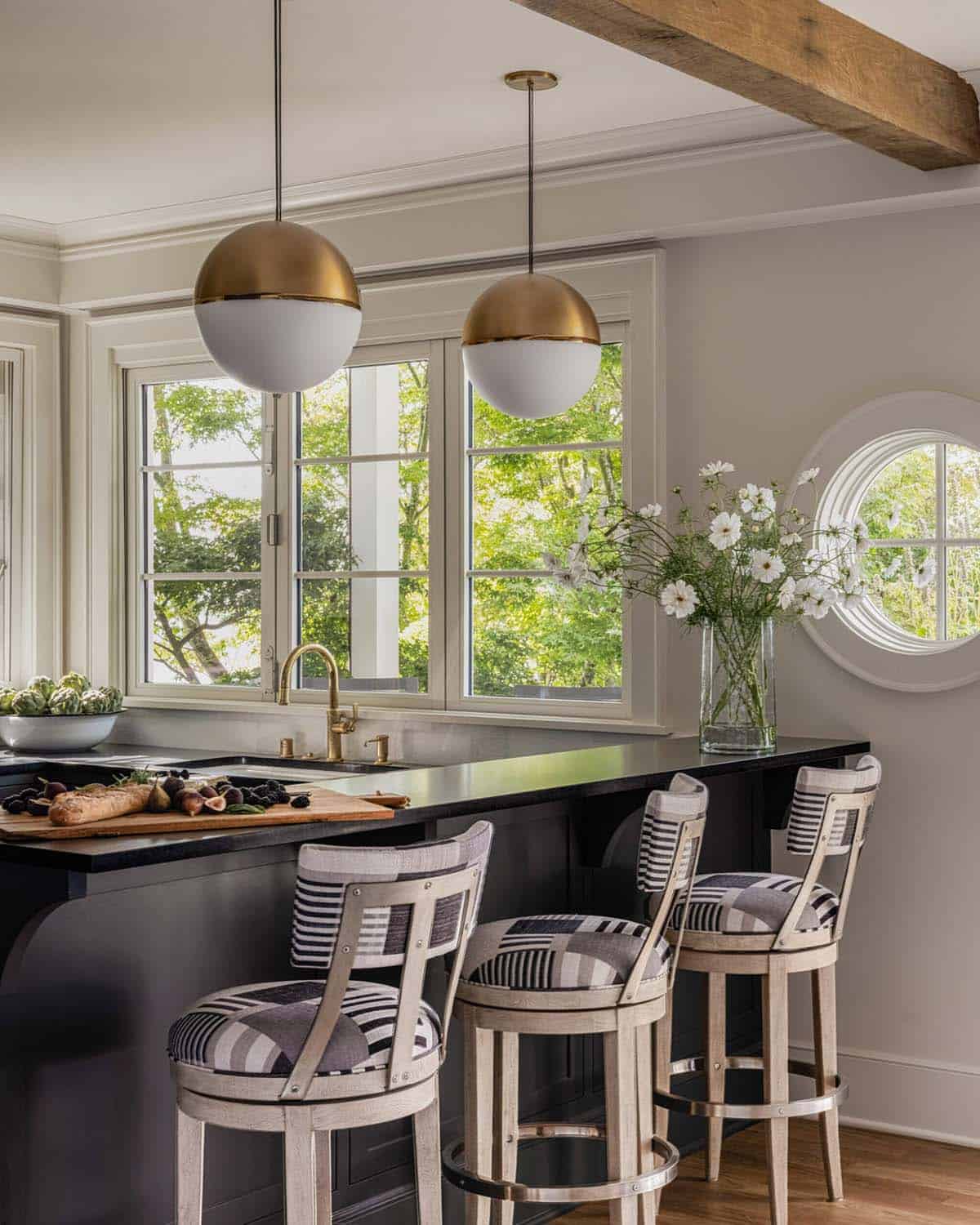 Kitchen island with striped barstools, gold globe pendants, porthole window, and tree views through casement windows