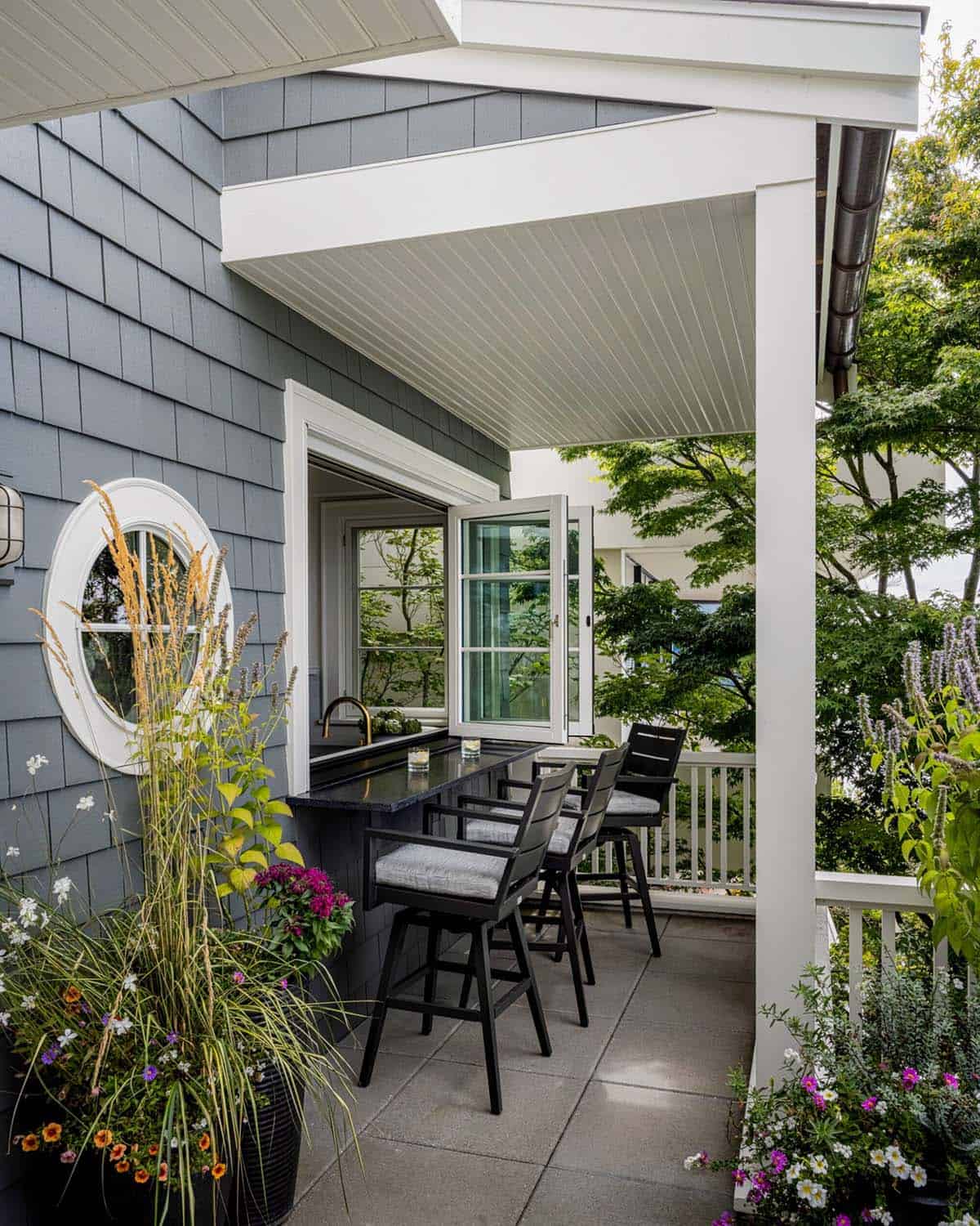 Exterior kitchen pass-through window bar with dark counter seating, porthole window, beadboard porch ceiling, and flowering planters