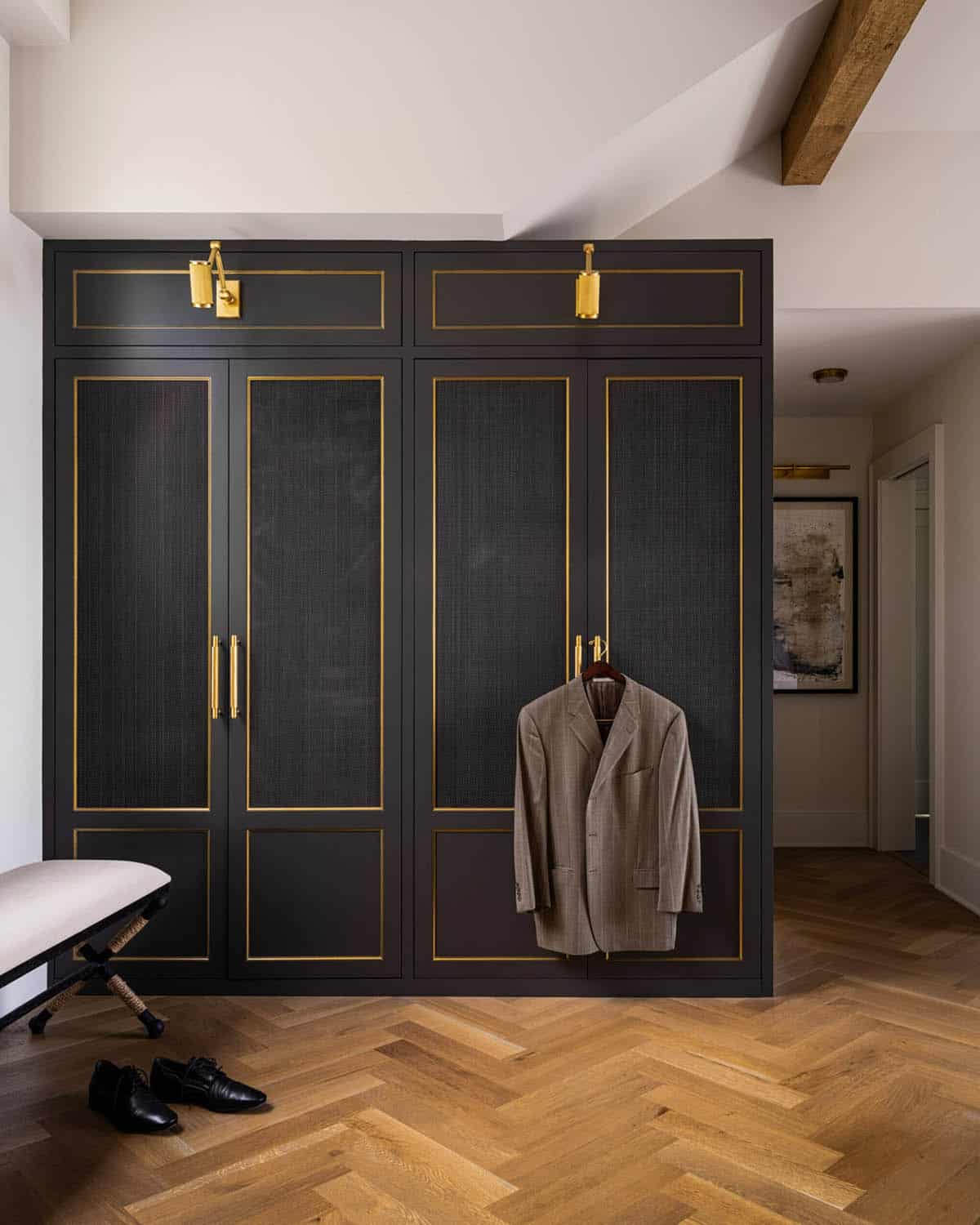 Full-wall dark wardrobe with grasscloth panel doors, brass inlay trim, picture lights, and herringbone oak floor