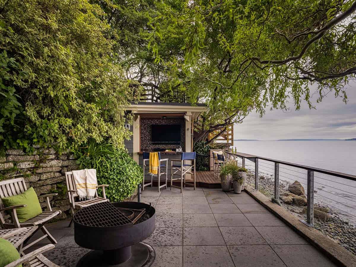 Waterfront stone terrace with fire pit, original boathouse bar, lush overhanging trees, and Puget Sound beyond