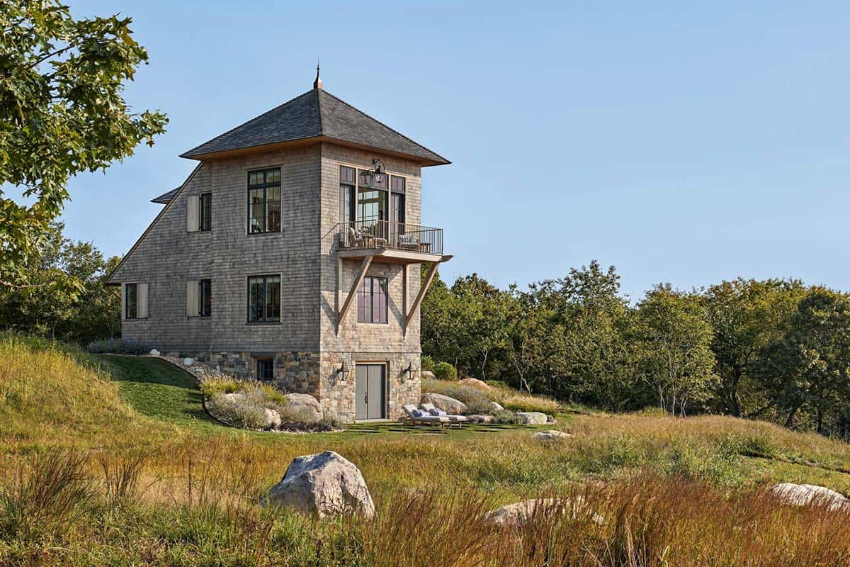 Three-story shingle and stone tower house perched on hillside surrounded by wild grasses