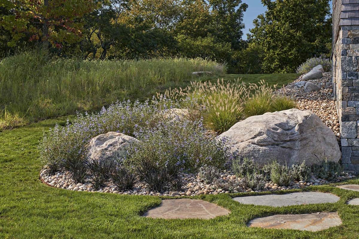 Native perennial garden with boulders, ornamental grasses, and river stone mulch beside stone foundation