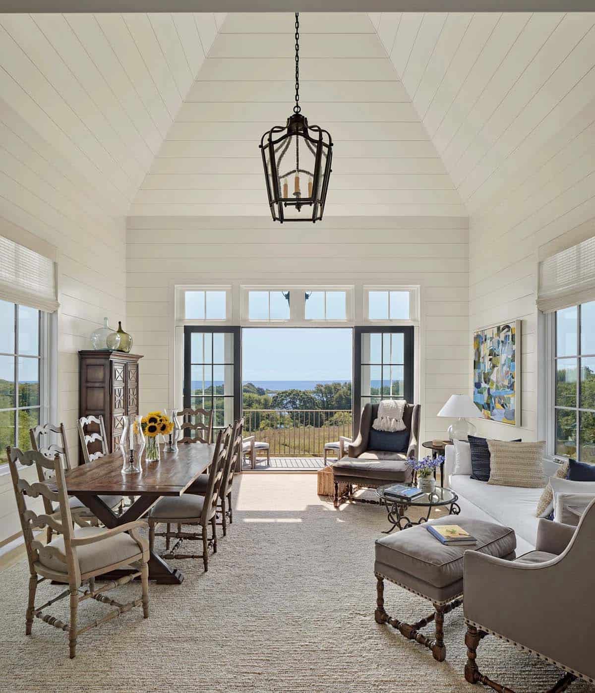 Open living and dining room with shiplap ceiling, lantern pendant, antique table, and ocean balcony beyond