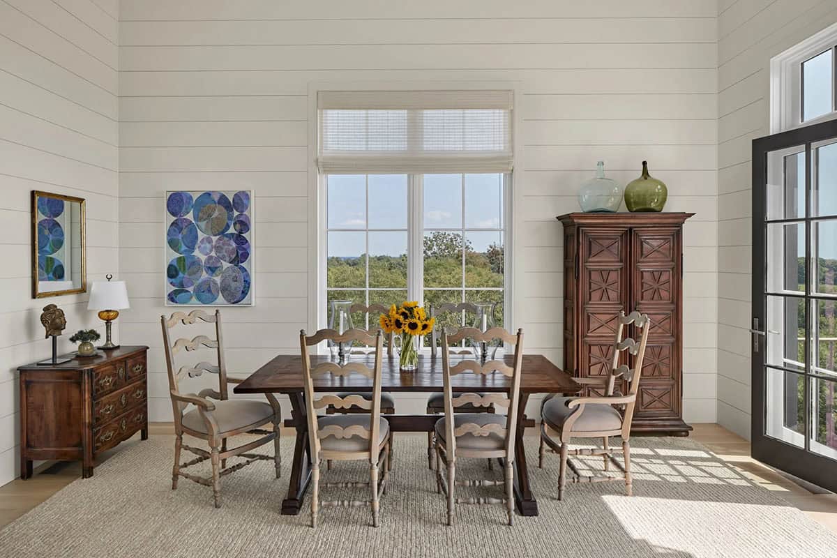 Bright dining room with shiplap walls, dark trestle table, ladder-back chairs, and antique armoire