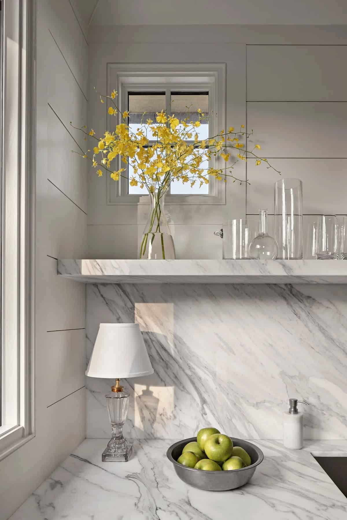 Kitchen detail of marble shelf and backsplash with yellow orchid stems and bowl of green apples
