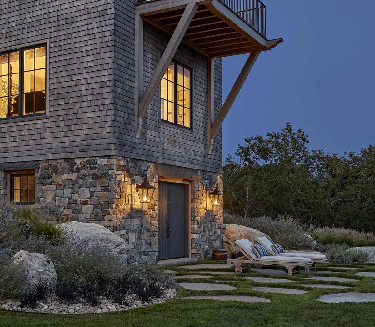 Tower house exterior at dusk with glowing windows, stone base, lantern sconces, and lounge chairs
