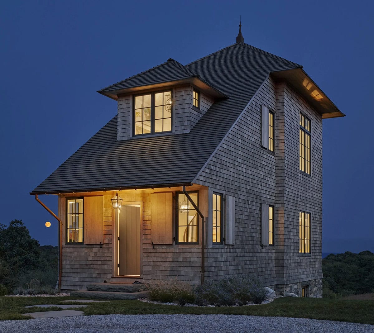 Tower house exterior at dusk with glowing windows, stone base, lantern sconces, and lounge chairs