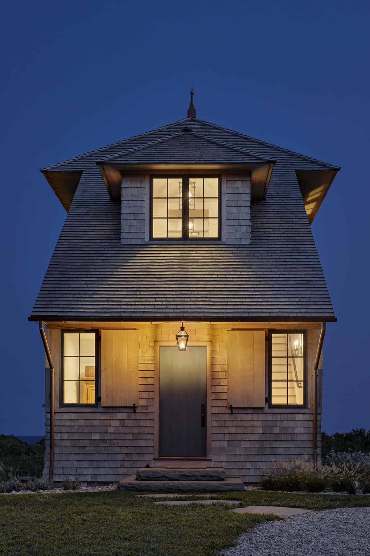 Tower house exterior at dusk with glowing windows, stone base, lantern sconces, and lounge chairs