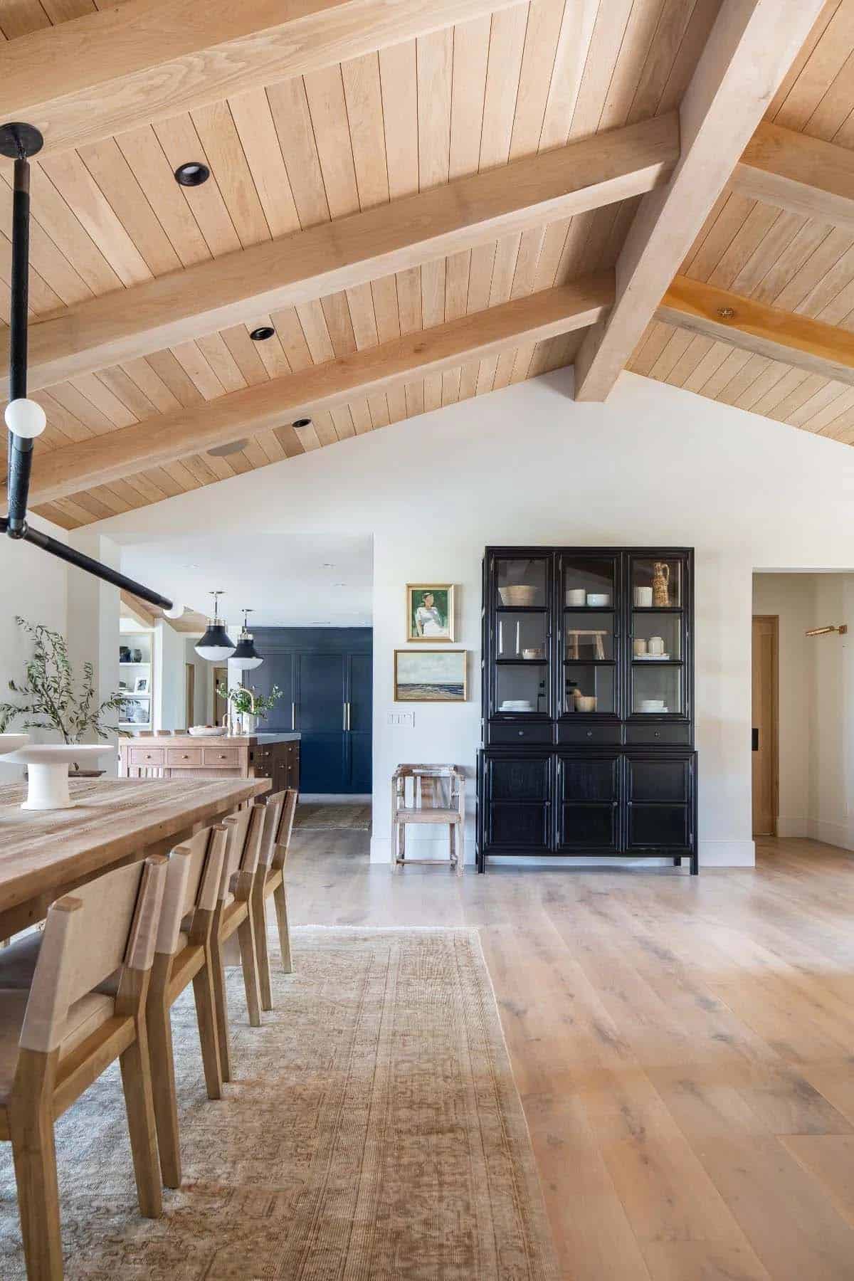 Dining area with vaulted wood beam ceiling, rustic dining table, and open view into navy blue kitchen
