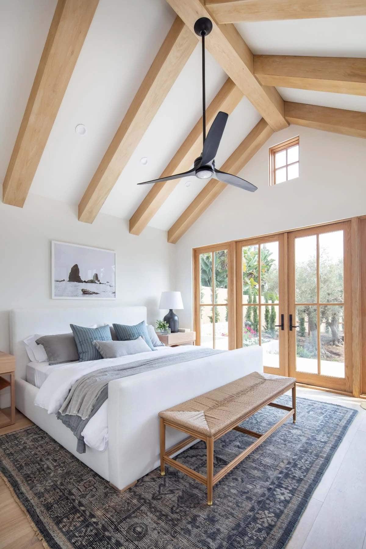 Primary bedroom with wood beam vaulted ceiling, white upholstered bed, and French doors opening to garden