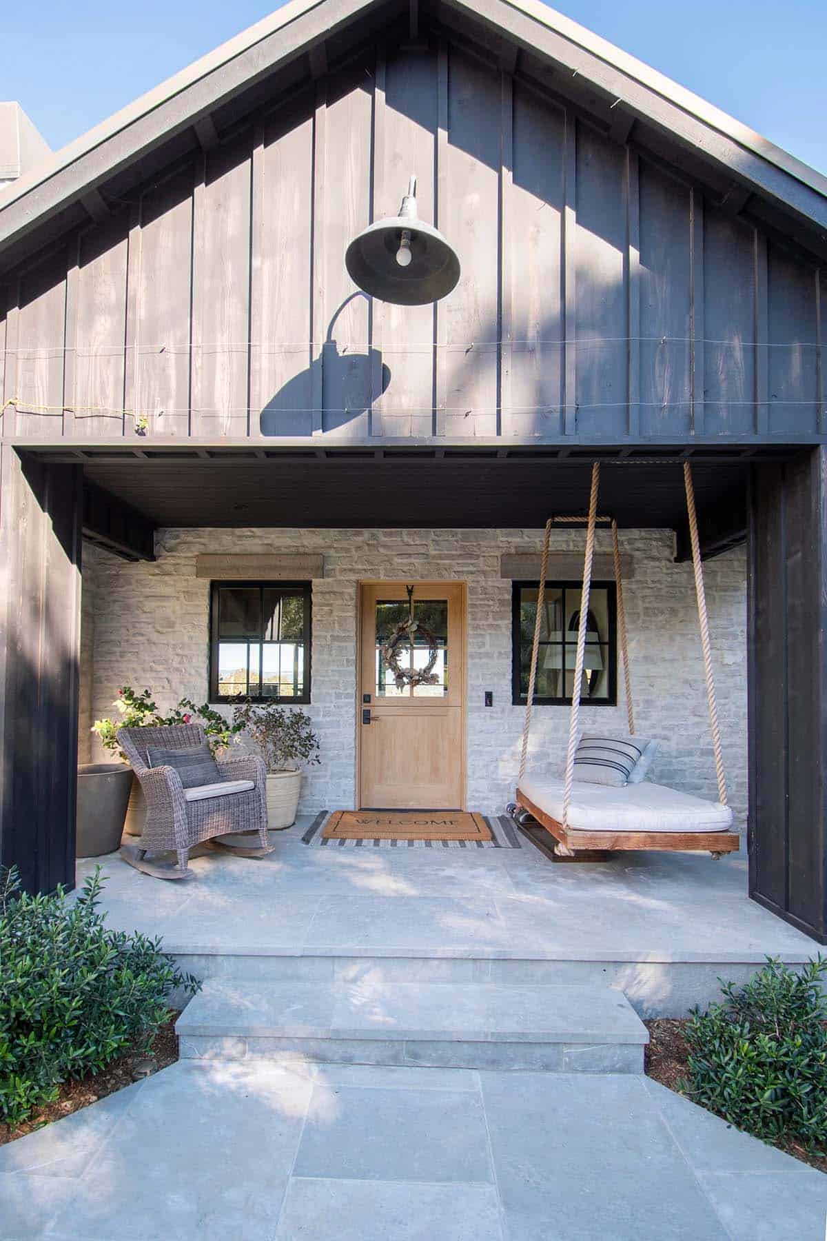 Front porch of a coastal home with dark board-and-batten siding, stone facade, rope porch swing, and rocking chair