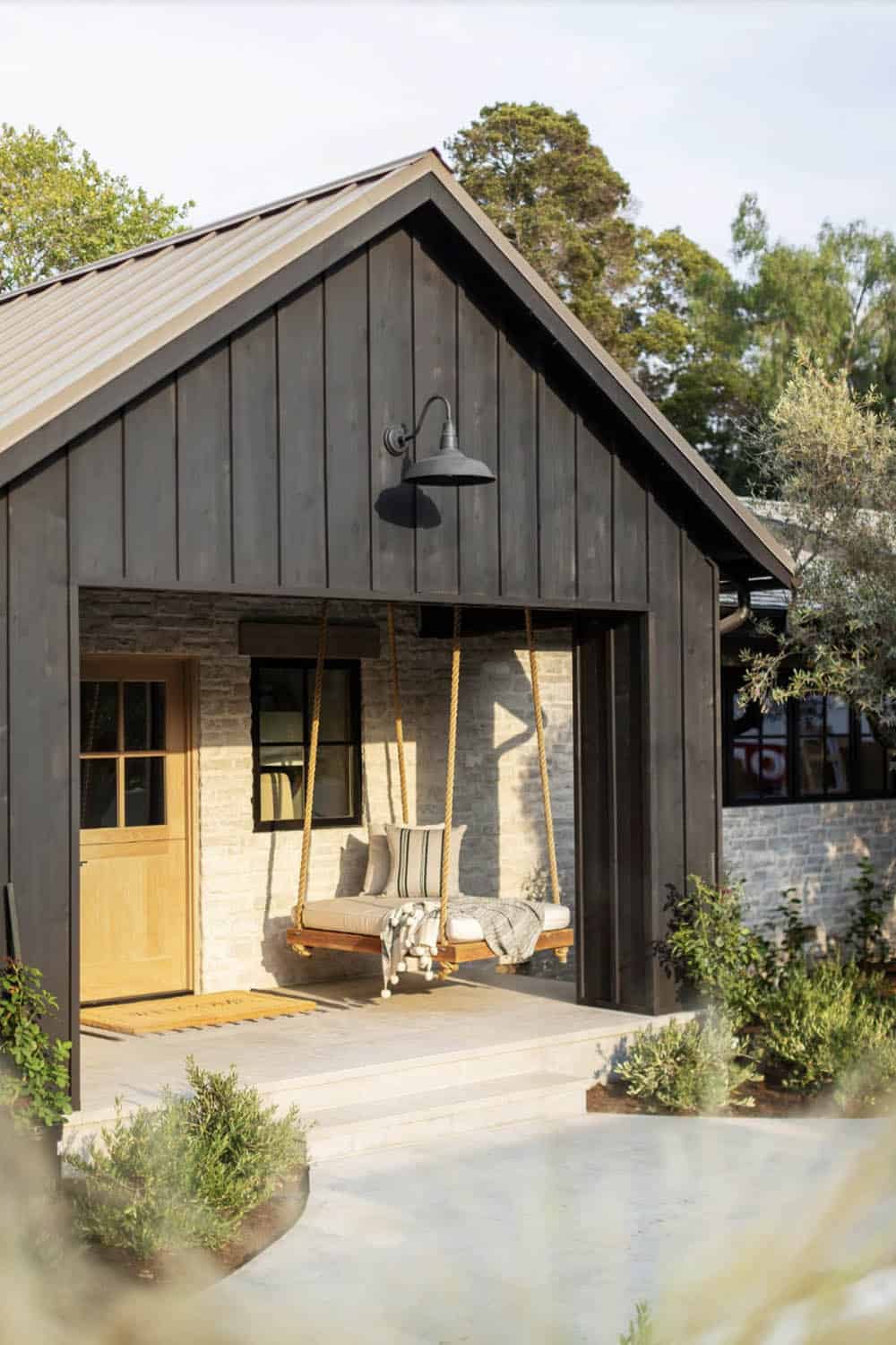 Angled exterior view of a dark board-and-batten coastal home with rope porch swing and stone steps