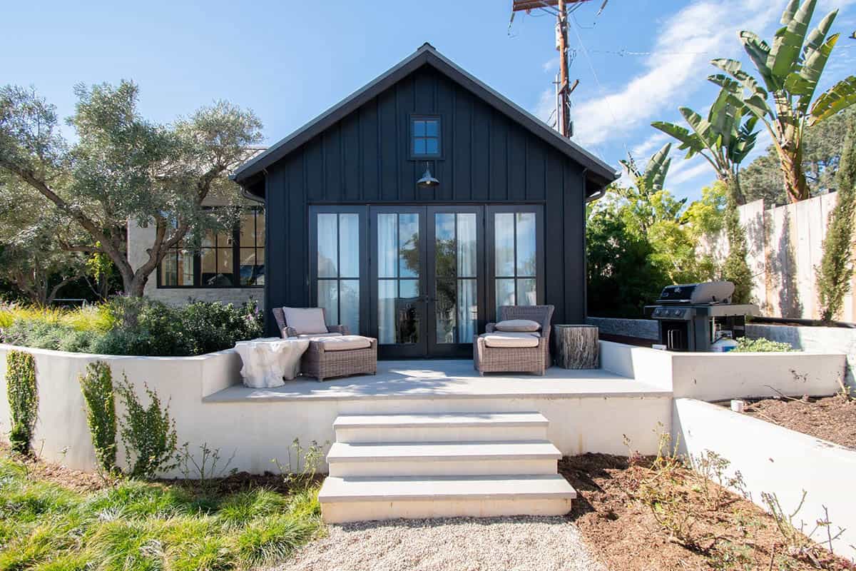 Rear exterior of a dark board-and-batten guest house with French doors, concrete patio, and wicker seating