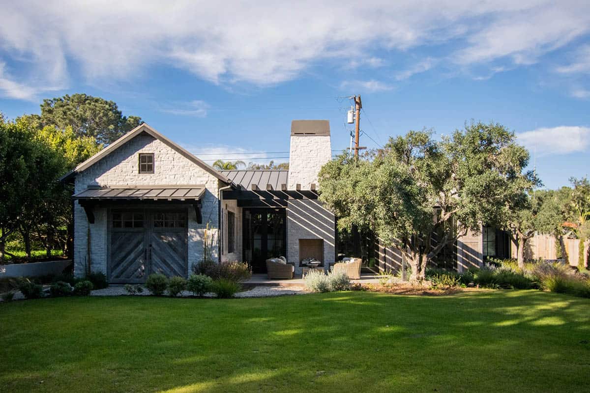 Rear exterior of a traditional coastal home with stone facade, metal roof, rustic barn doors, and mature olive trees