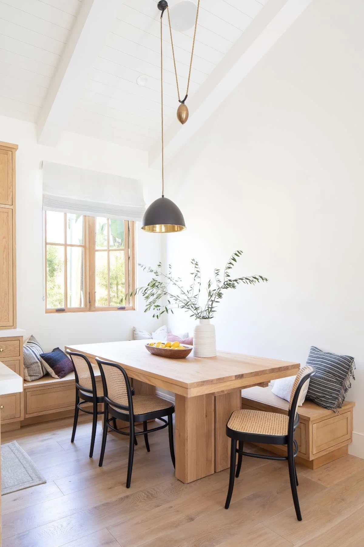 Bright kitchen breakfast nook with built-in oak banquette, cane chairs, wood pedestal table, and black dome pendant
