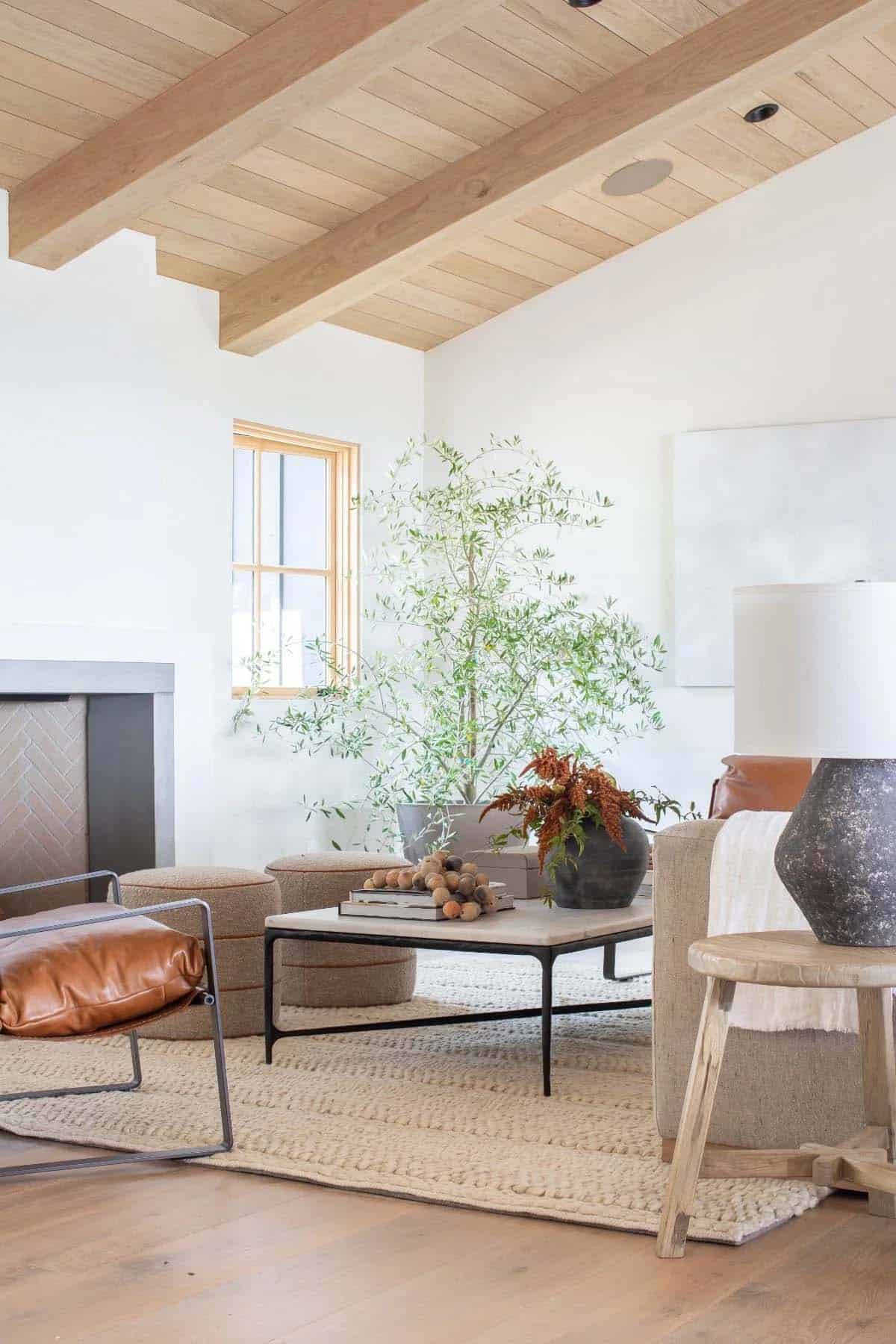 Living room corner with wood beam ceiling, leather chair, stone fireplace, ottomans, and large potted olive tree