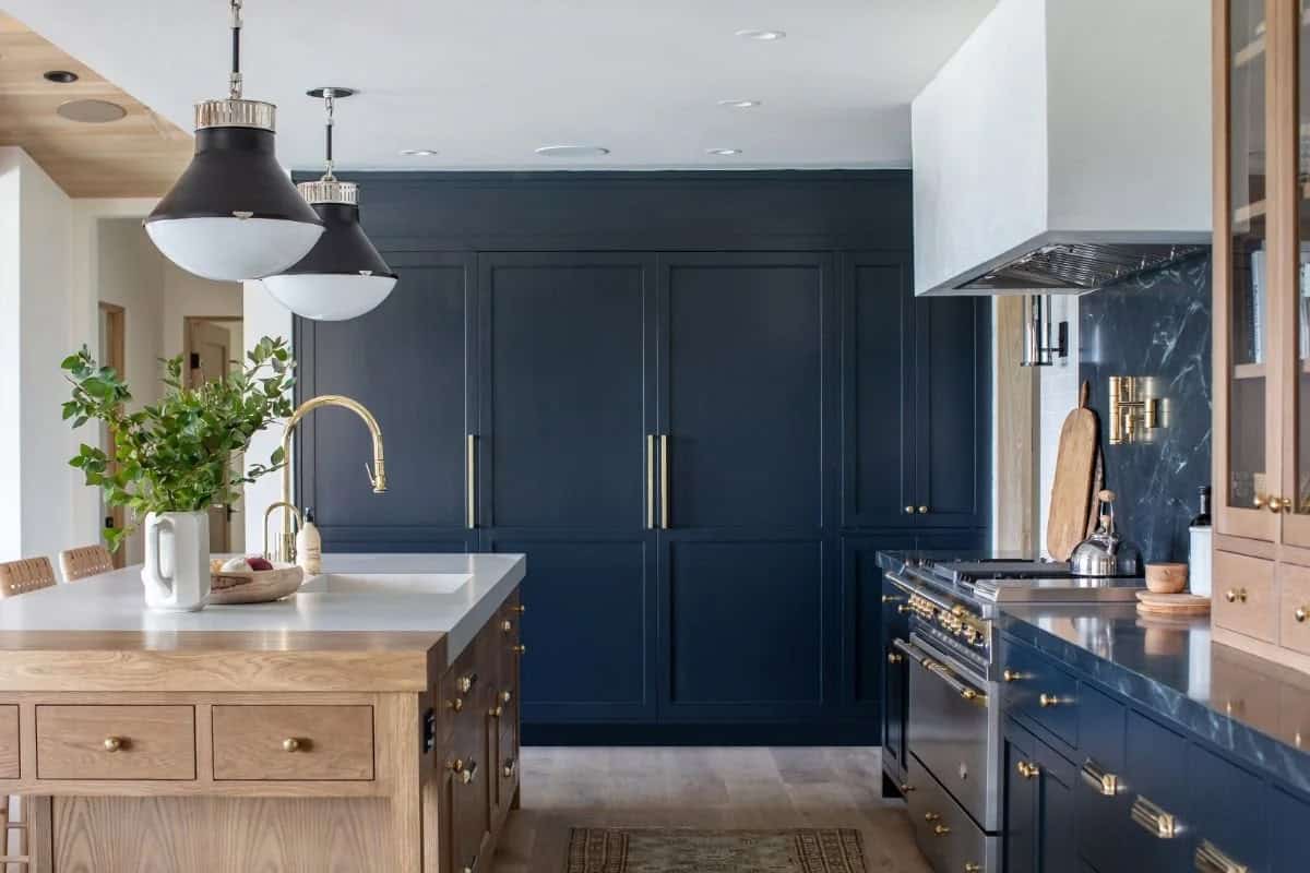 Navy blue kitchen with brass hardware, freestanding oak island, dark marble backsplash, and black dome pendants