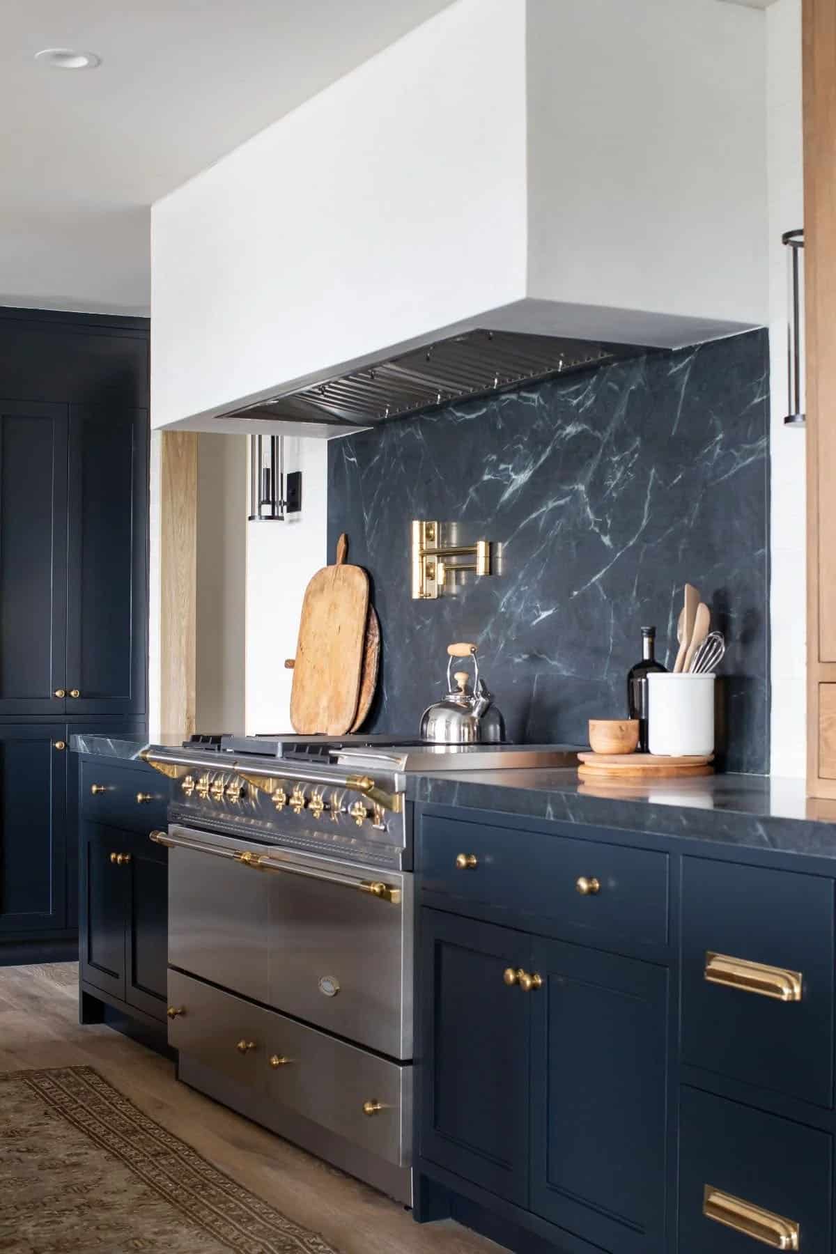 Close-up of navy kitchen range with dark marble slab backsplash, professional stainless range, and brass pot filler