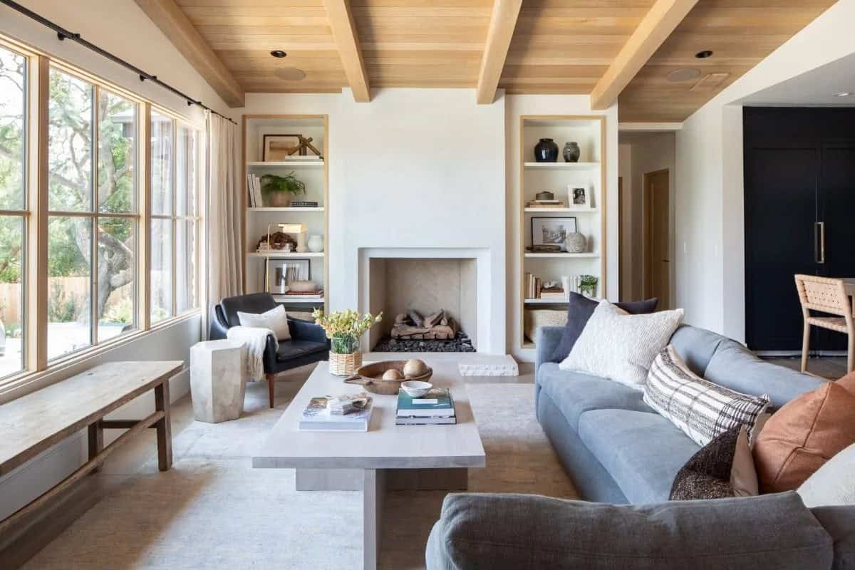 Living room with wood beam ceiling, stone fireplace, flanking built-in shelves, gray sofa, and large windows