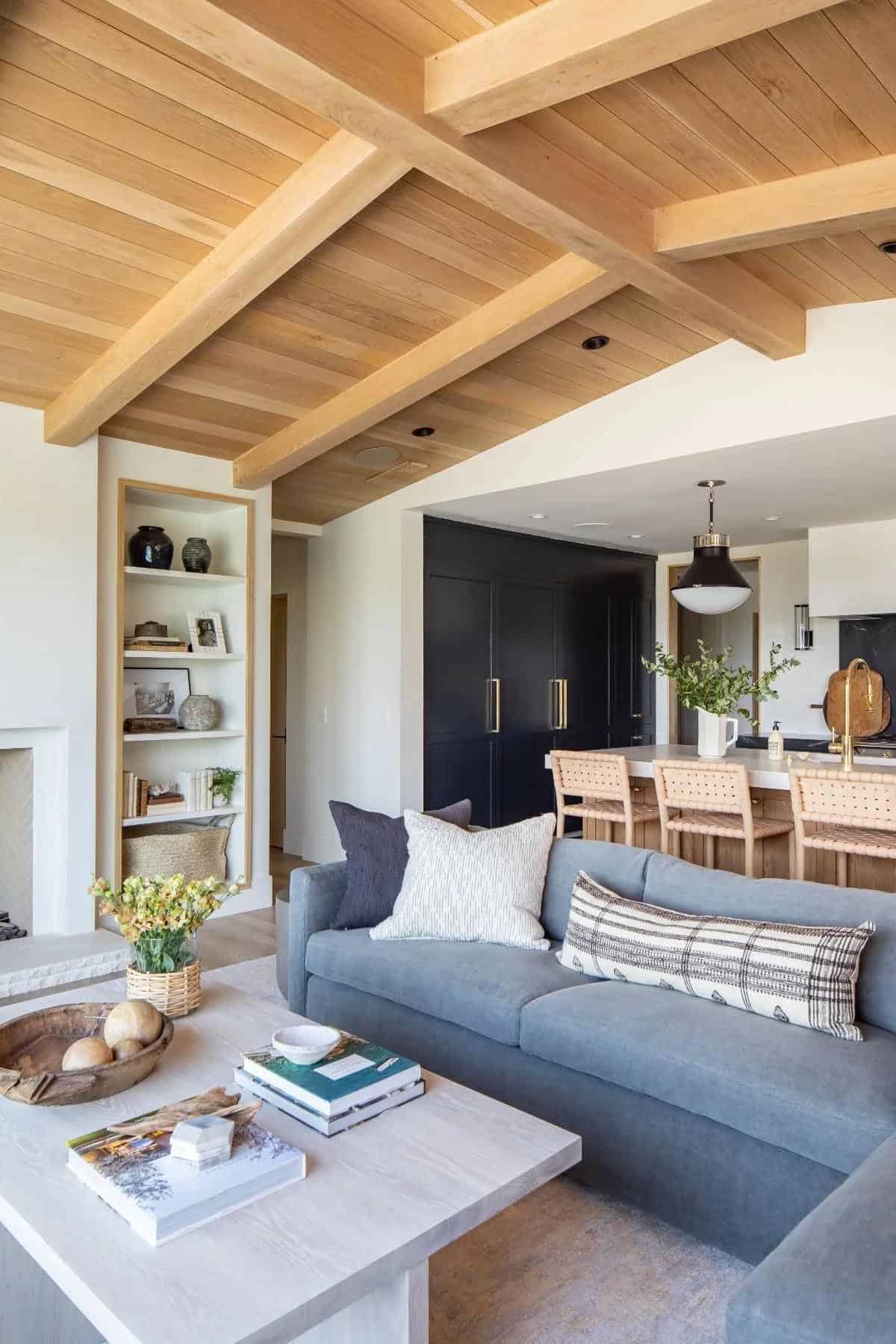 Open-plan living room with wood beam ceiling, gray sofa, built-in shelves, and view into navy blue kitchen