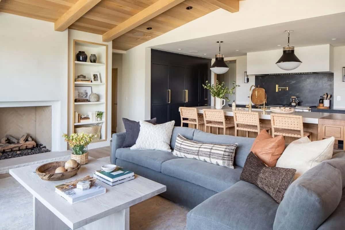 Coastal living room with gray sectional, stone fireplace, wood beam ceiling, and open view to navy kitchen beyond