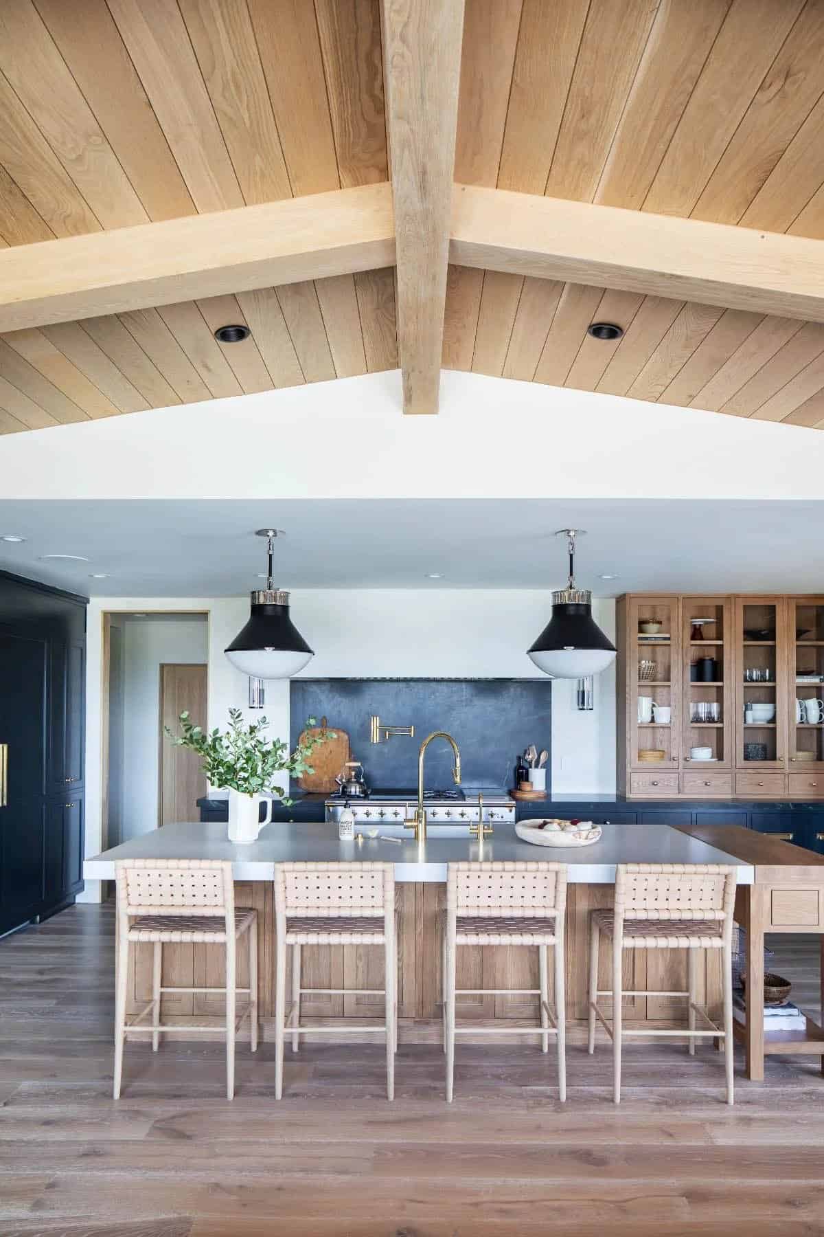 Kitchen island with woven bar stools under wood shiplap vaulted ceiling, navy cabinets, and oak hutch beyond