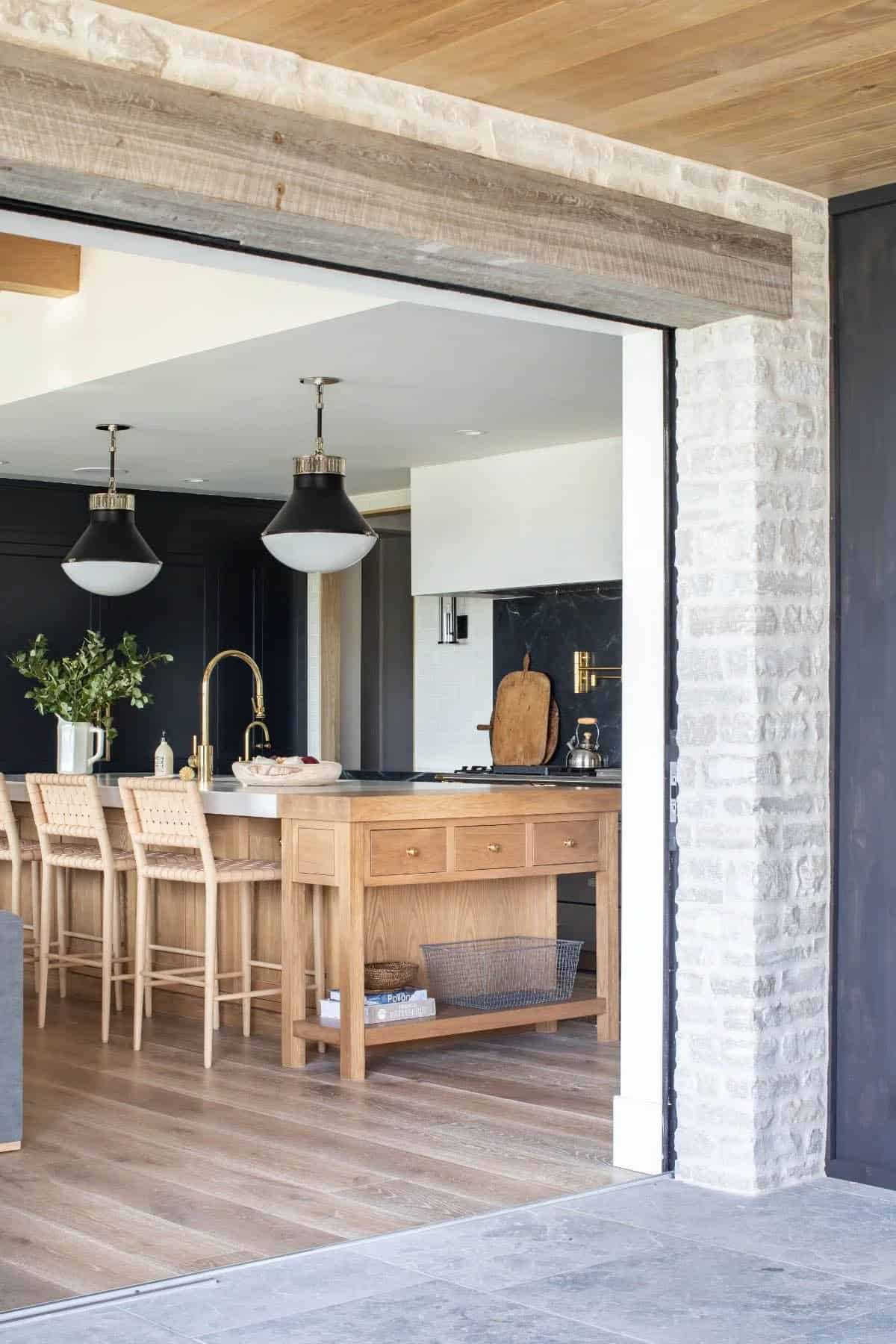 View into coastal kitchen through stone and reclaimed wood opening with oak island and woven bar stools