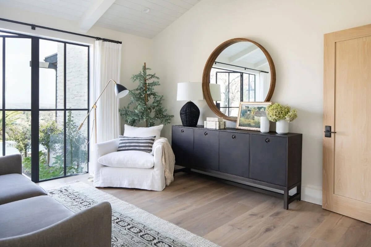 Sitting room with black sideboard, round wood mirror, white armchair, and black-framed glass doors