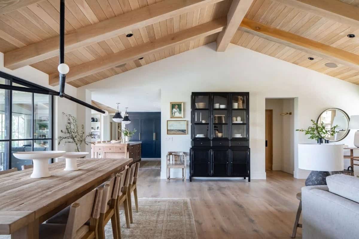 Open-plan dining room with vaulted wood beam ceiling, rustic table, black cabinet, and view to navy kitchen