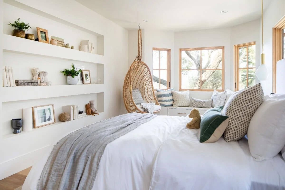 Children's bedroom with built-in white shelves, rattan hanging chair, window seat, and white bedding