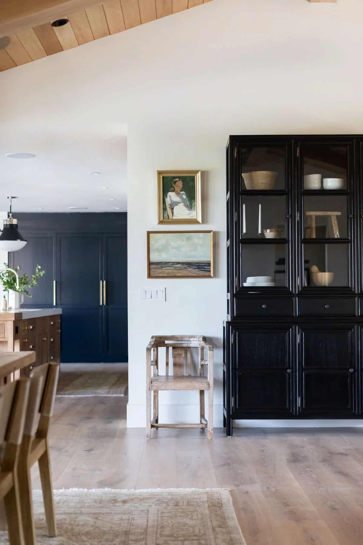 dining room, black glass-front cabinet, gold-framed coastal artwork, open plan to navy kitchen beyond