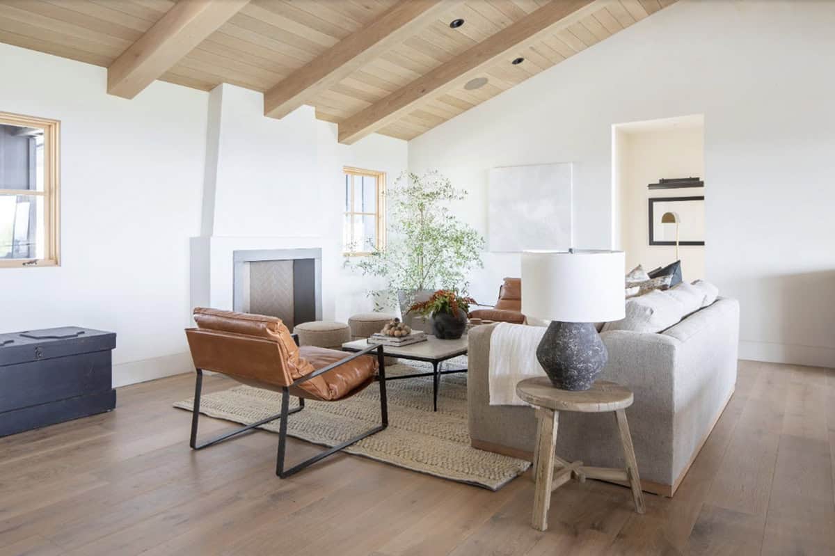 Living room corner with wood beam ceiling, leather chair, stone fireplace, ottomans, and large potted olive tree