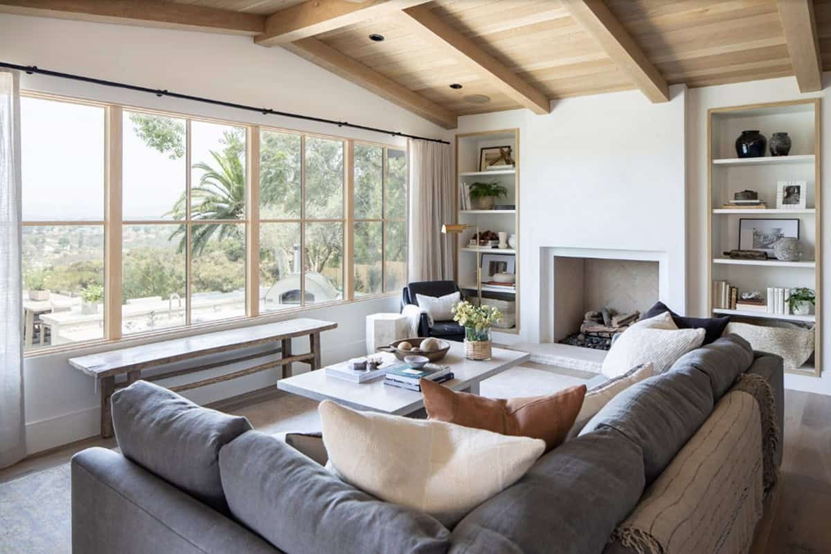 Living room with wood beam ceiling, stone fireplace, flanking built-in shelves, gray sofa, and large windows