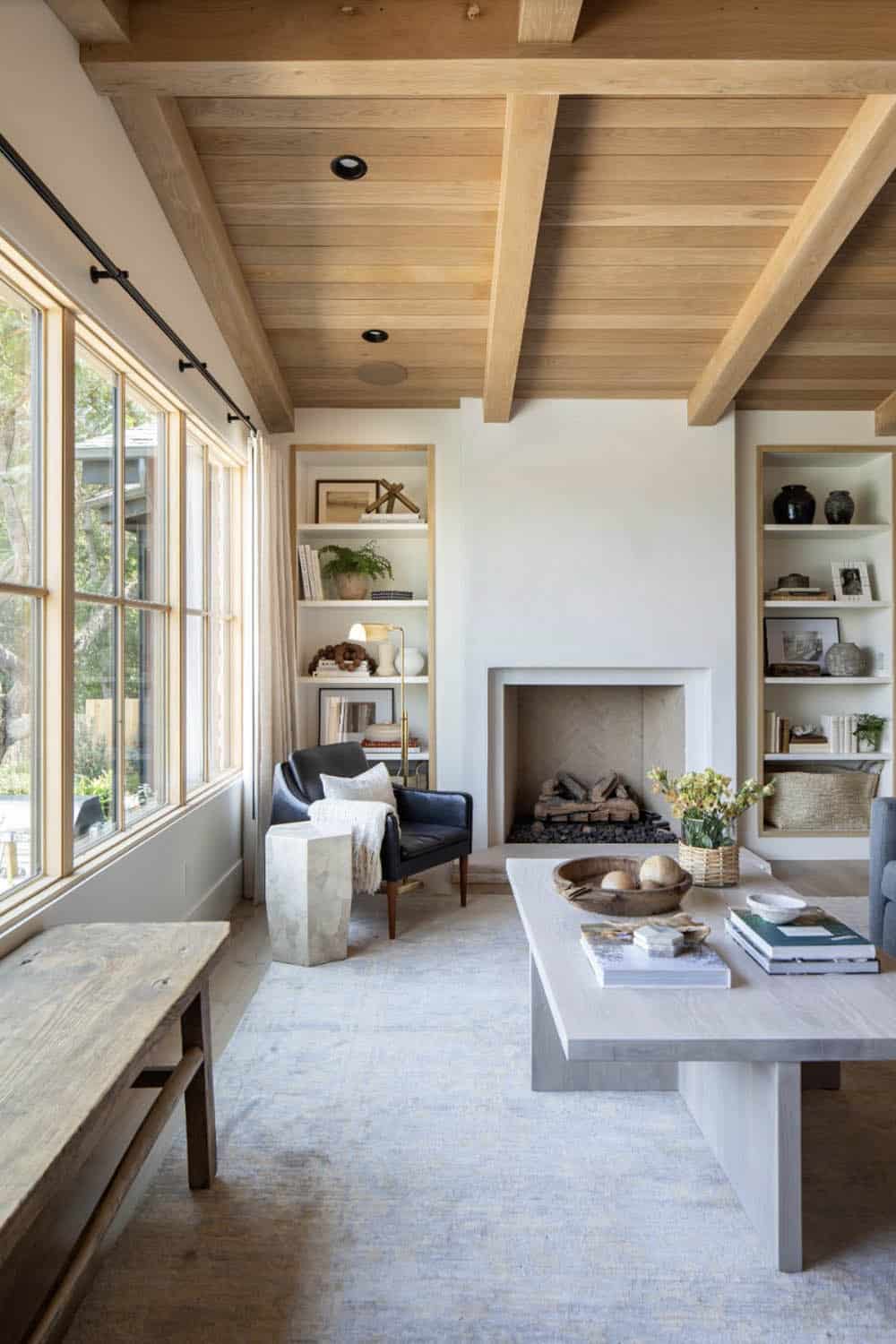 Living room with wood beam ceiling, stone fireplace, flanking built-in shelves, gray sofa, and large windows
