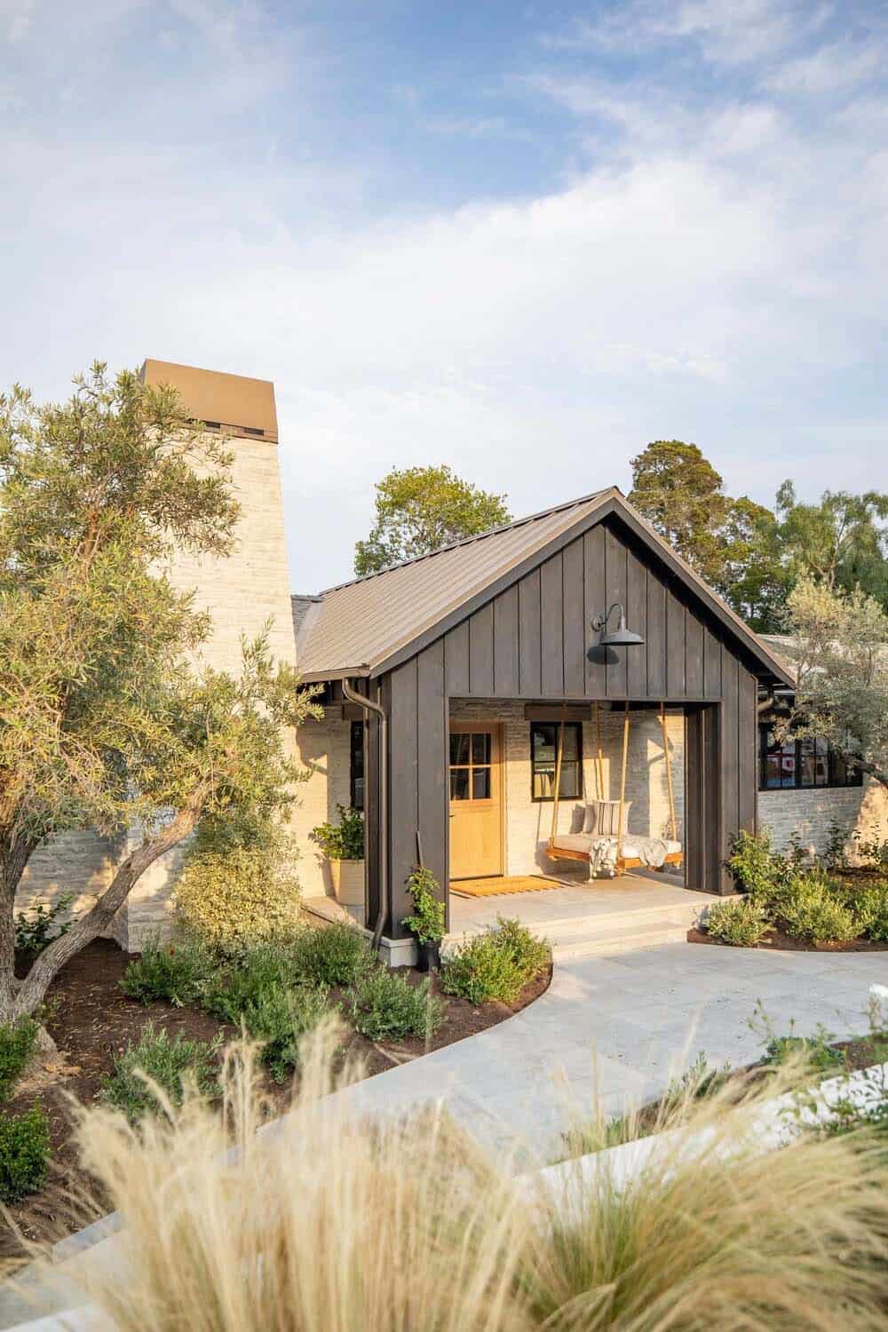 Front porch of a coastal home with dark board-and-batten siding, stone facade, rope porch swing, and rocking chair