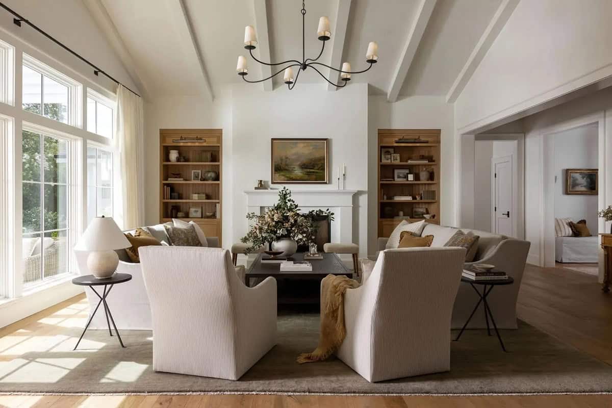 Bright living room with vaulted beamed ceiling, flanking oak built-ins, white fireplace, and cream armchairs