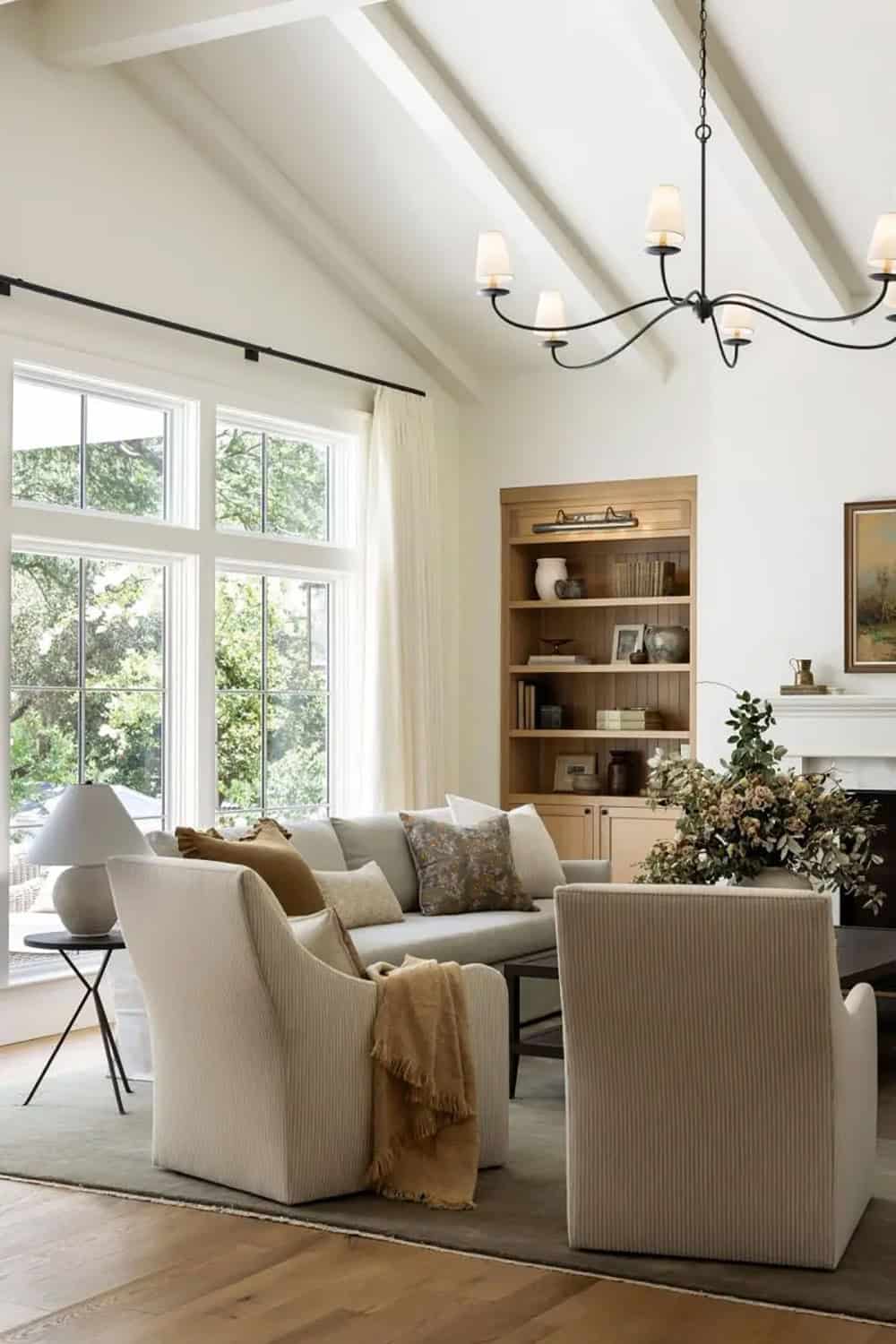 Bright living room with vaulted ceiling, cream armchairs, iron chandelier, and large oak built-in bookcase