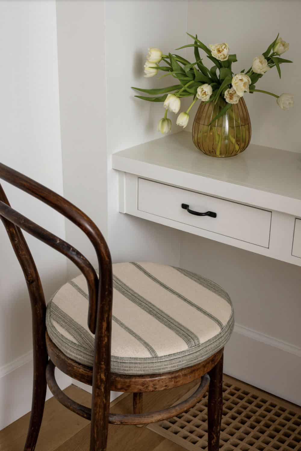 Close-up of bentwood chair with striped cushion beside white built-in desk with white tulips in amber vase
