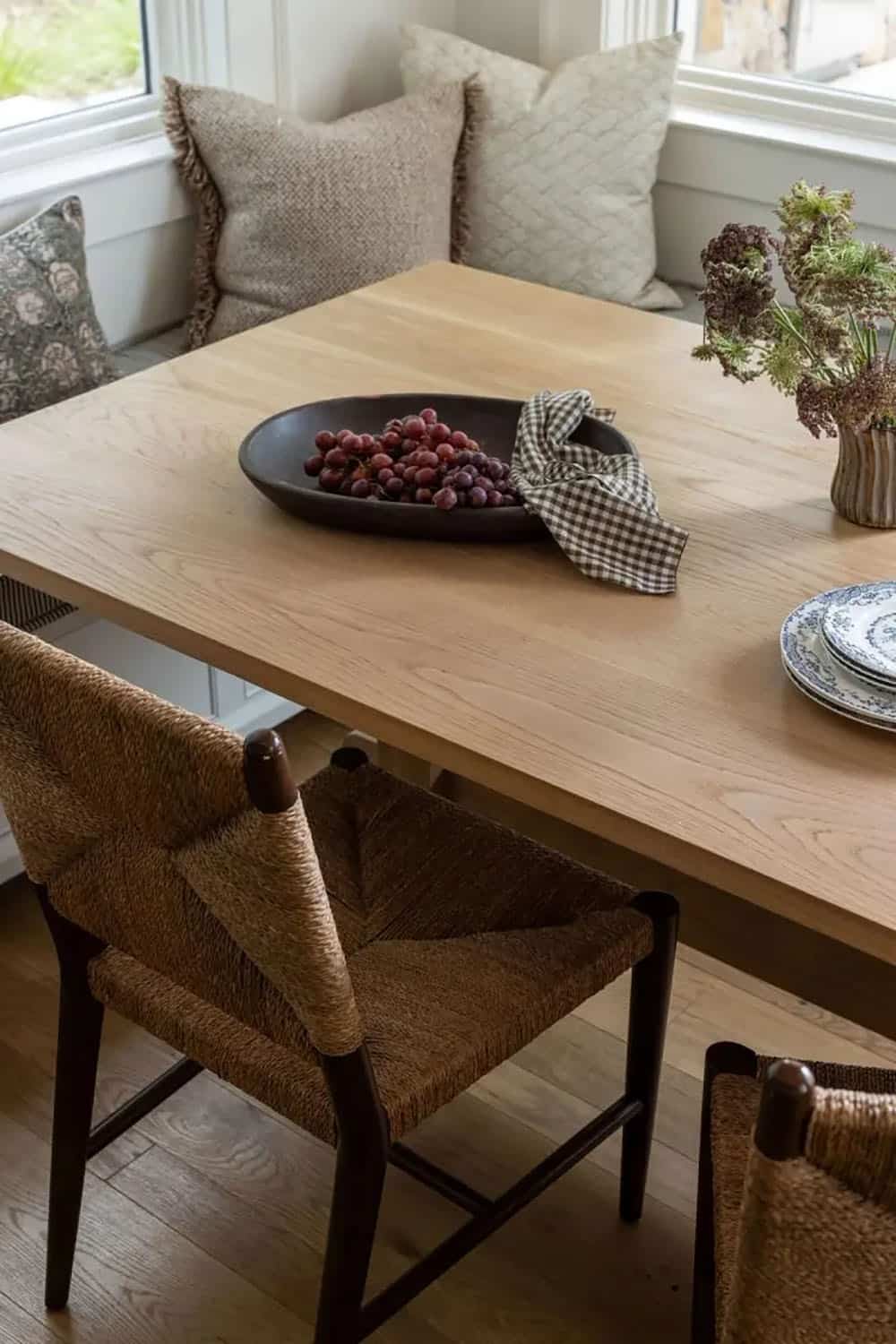 Close-up of oak breakfast nook table with woven rush chair, dark bowl of grapes, and gingham napkin