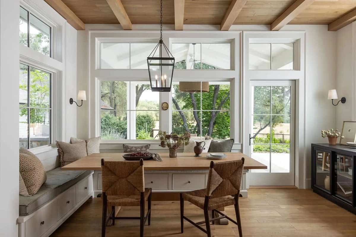 Sun-filled breakfast nook with wraparound window seat, oak ceiling beams, black lantern pendant, and garden views